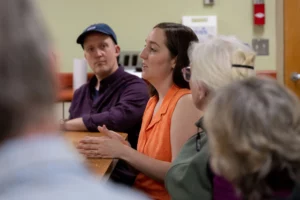 Mari Brunner, a woman with brown hair and wearing a sleeveless orange blouse, is a senior planner with the Keene community development department and is shown at a meeting speaking with clean energy and community power advocates on April 5, 2023, at City Hall in Keene, N.H.